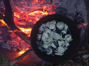 Auf dem Lagerfeuer steht ein gusseiserner Topf, darin werden Zwiebeln gebraten