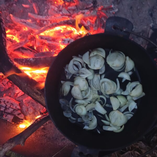 Auf dem Lagerfeuer steht ein gusseiserner Topf, darin werden Zwiebeln gebraten